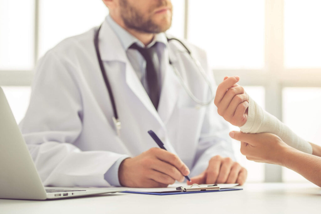 Doctor examining a patient’s bandaged wrist while taking notes at a desk.